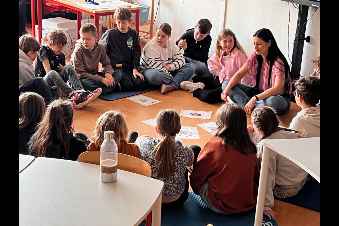Eine Schulklasse sitzt im Kreis auf dem Boden, in der Mitte liegen Karten und Tablets.