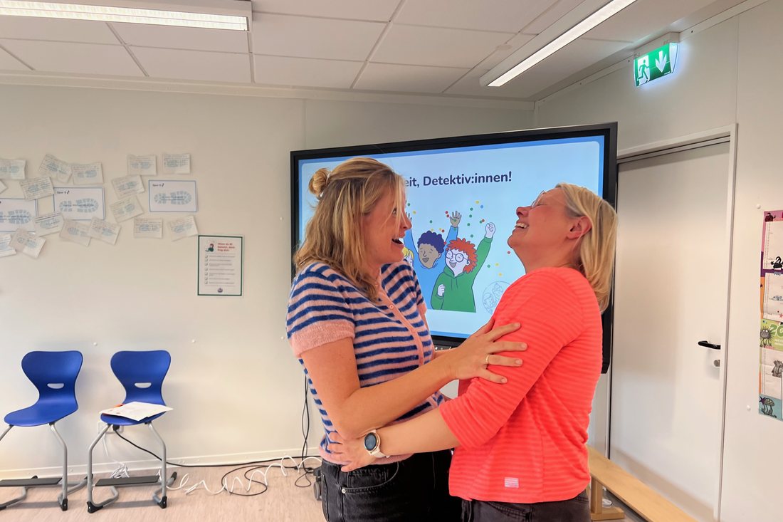 Zwei Lehrerinnen lachen und halten einander an den Armen. Hinter ihnen zeigt der Bildschirm die Abschlussfolie mit jubelnden Kindern und Konfetti.