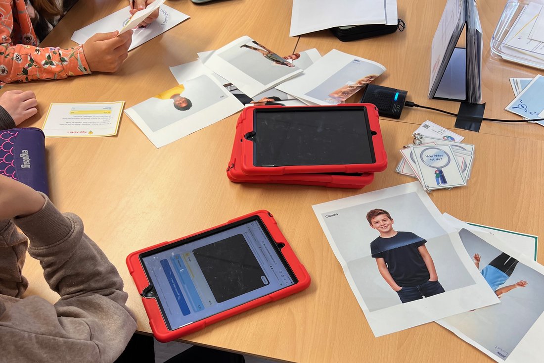 Gruppentisch im Klassenraum: zwei Tablets in roten Schutzhüllen, daneben ein kleines schwarzes Mikrofon mit leuchtender Betriebsanzeige, dessen Kabel mit schwarzem Klebeband auf der Tischplatte fixiert ist. Am Rand der unteren Hülle steckt eine aufsteckbare Weitwinkel-Linse vor der Kamera.